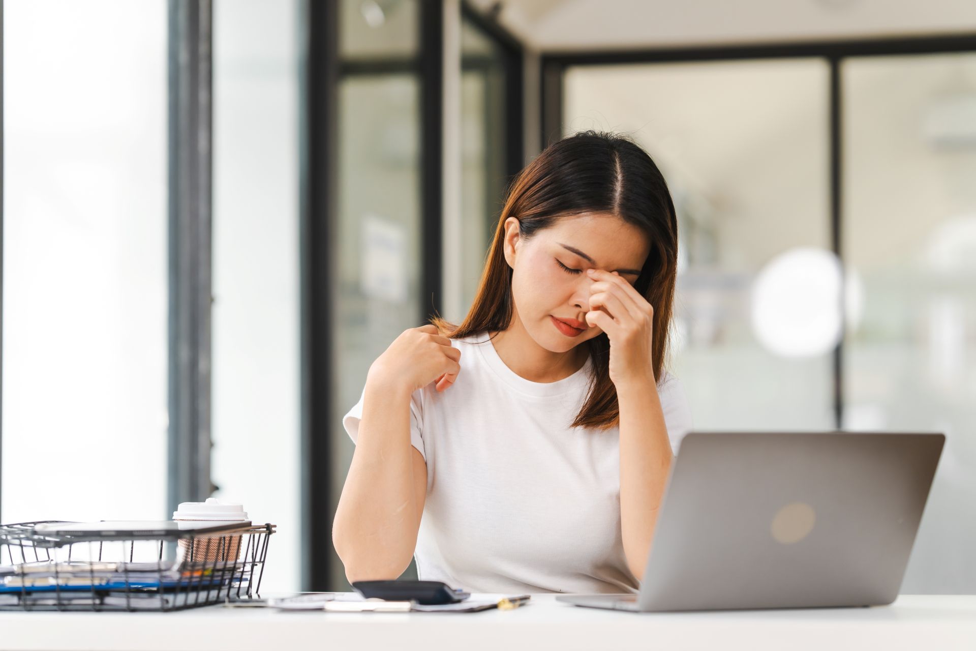 Frau sitzt im Buero am Laptop und reibt sich die Augen vor Stress, Symbol fuer psychische Belastung und die Bedeutung von Arbeitsschutzberatung