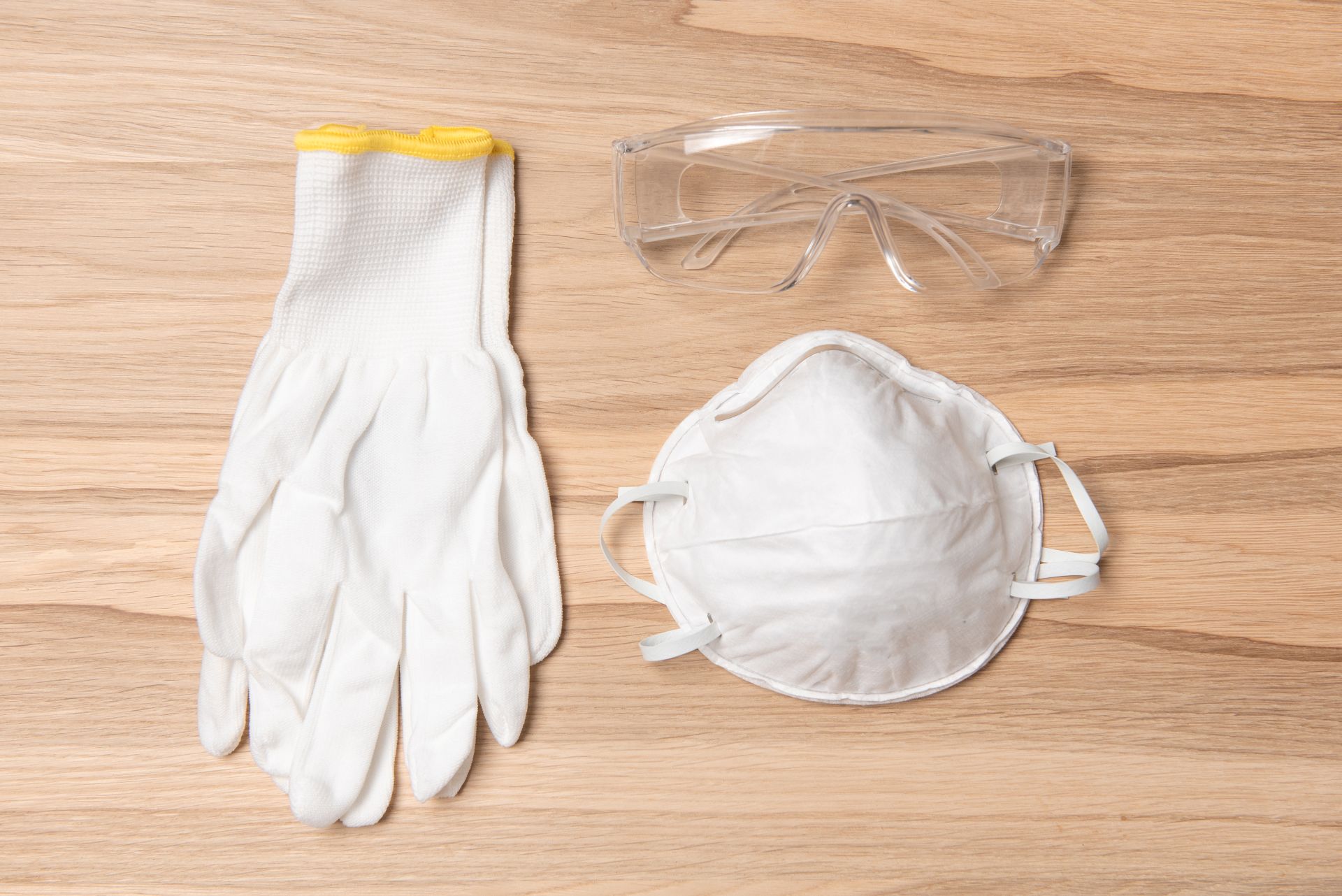 Weisse Handschuhe, Atemschutzmaske und Schutzbrille liegen auf einem Holztisch, Beispiel fuer grundlegende Arbeitsschutzmassnahmen